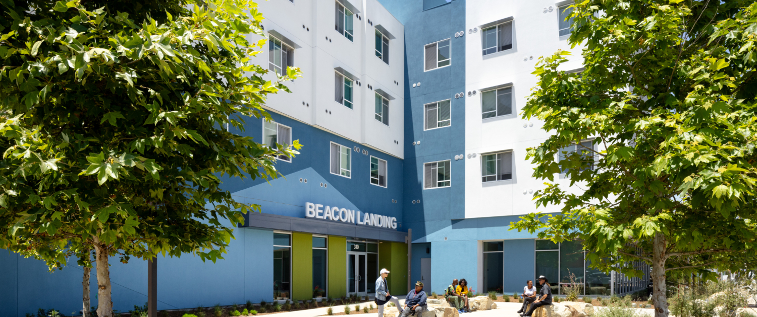 Exterior of building with white and blue facade. Beacon Landing site sign is centered above bright green door with angeled blue pattern in background. Residents site out front on large landscape rocks engaging with each other.