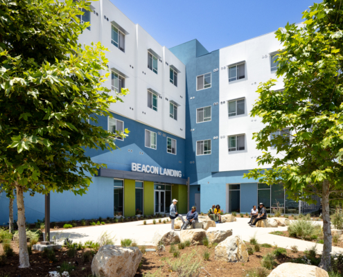 Exterior of building with white and blue facade. Beacon Landing site sign is centered above bright green door with angeled blue pattern in background. Residents site out front on large landscape rocks engaging with each other.