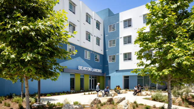 Exterior of building with white and blue facade. Beacon Landing site sign is centered above bright green door with angeled blue pattern in background. Residents site out front on large landscape rocks engaging with each other.