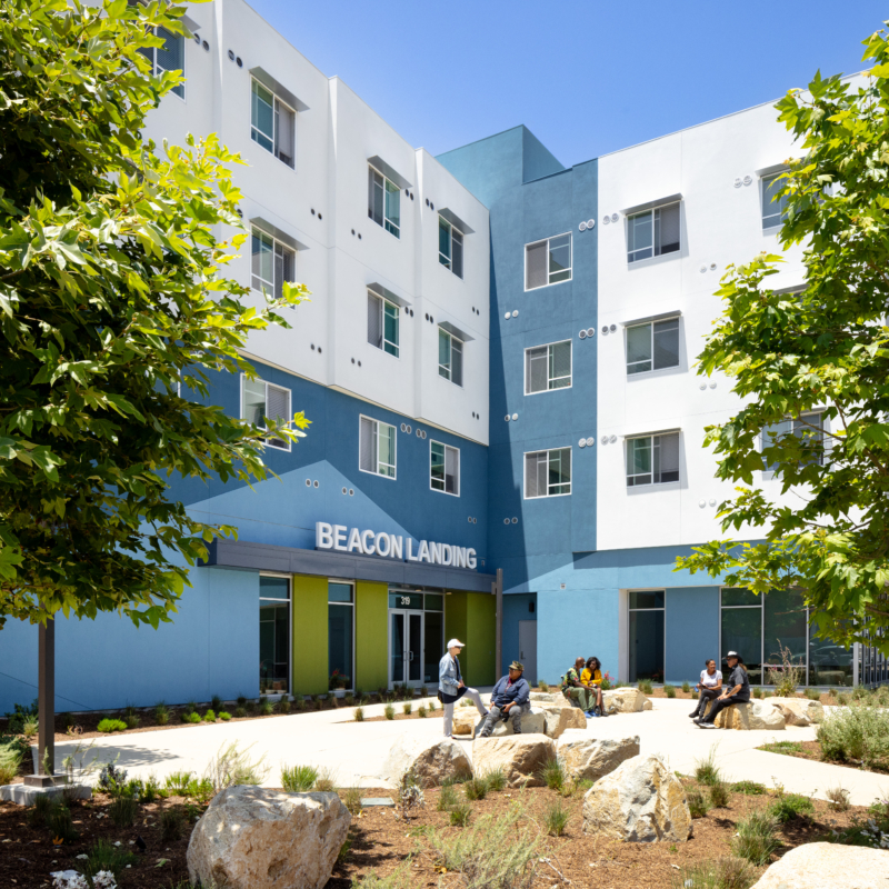 Exterior of building with white and blue facade. Beacon Landing site sign is centered above bright green door with angeled blue pattern in background. Residents site out front on large landscape rocks engaging with each other.
