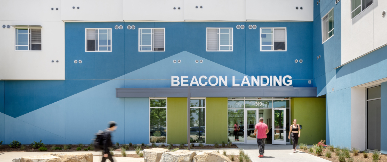 Front entrance of building with Beacon Landing sign in front of angled pattern featuring light blue and line green paint. Residents walking into building.