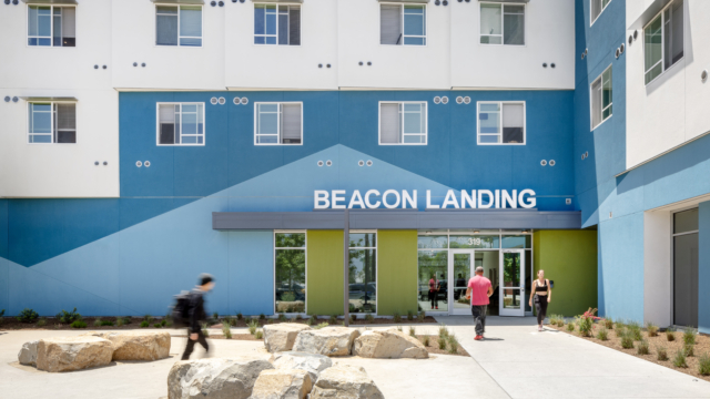 Front entrance of building with Beacon Landing sign in front of angled pattern featuring light blue and line green paint. Residents walking into building.