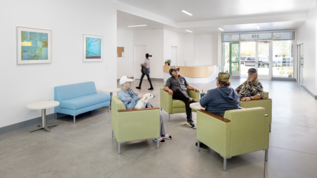 Interior lobby where residents are gathered talking. Four central green chairs surround table.