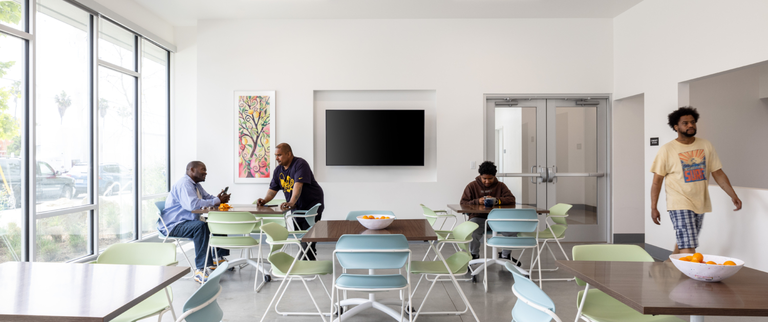Community room with residents chatting. Room is situated with five tables, each with four chairs. A television is off in the background. Colorful abstract art on wall.