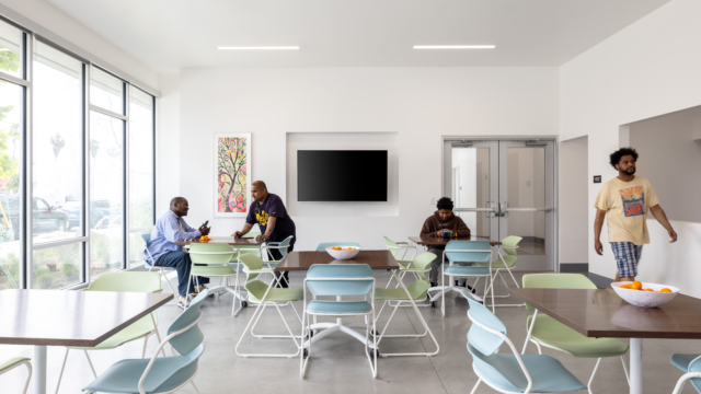 Community room with residents chatting. Room is situated with five tables, each with four chairs. A television is off in the background. Colorful abstract art on wall.