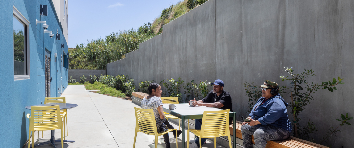 Three residents chatting in back of building in long courtyard. Concrete retaining wall in the back protects the property. Green simple landscaping creates a warm and inviting space.