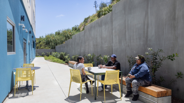 Three residents chatting in back of building in long courtyard. Concrete retaining wall in the back protects the property. Green simple landscaping creates a warm and inviting space.