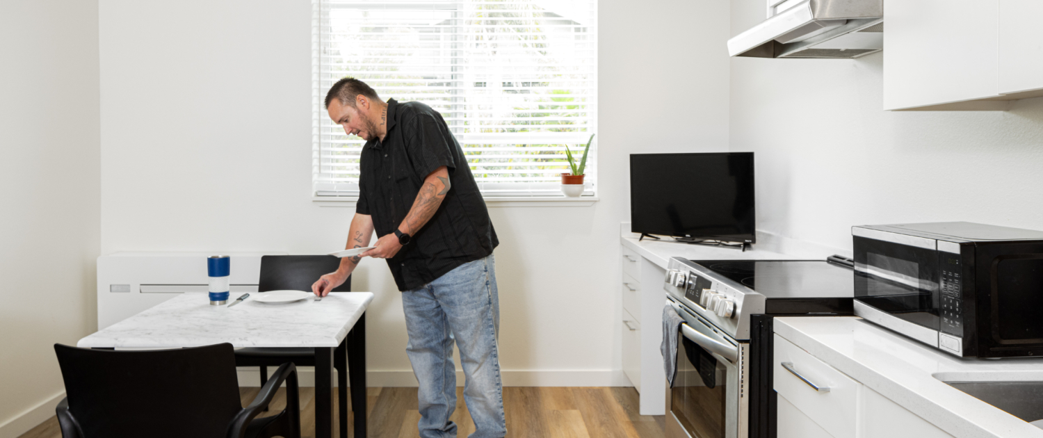 Picture of resident inside his dwelling. Resident is leaning over small dining room table to the left, with stove, countertop and storage to the right.