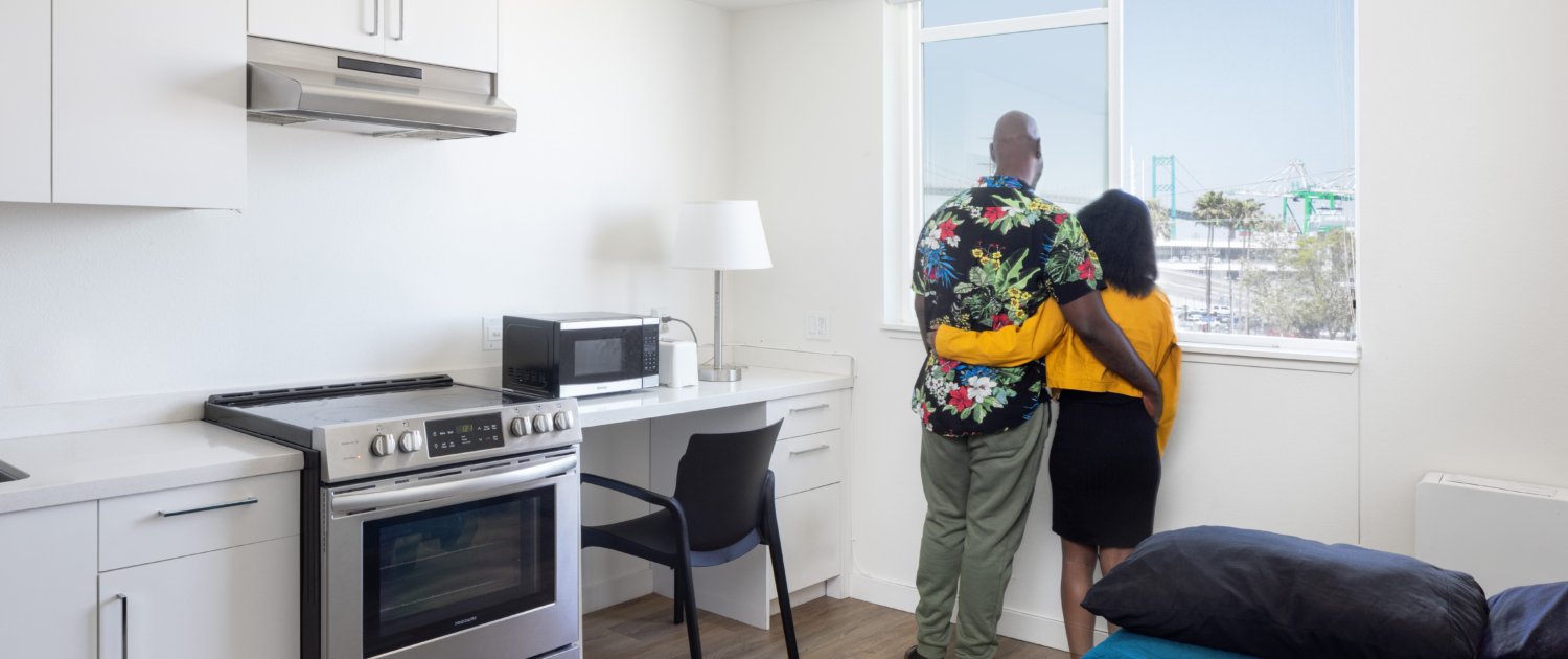 Two residents have their arms wrapped around each other as they look outside their unit window to a view of the Port of Los Angeles.