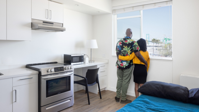 Two residents have their arms wrapped around each other as they look outside their unit window to a view of the Port of Los Angeles.