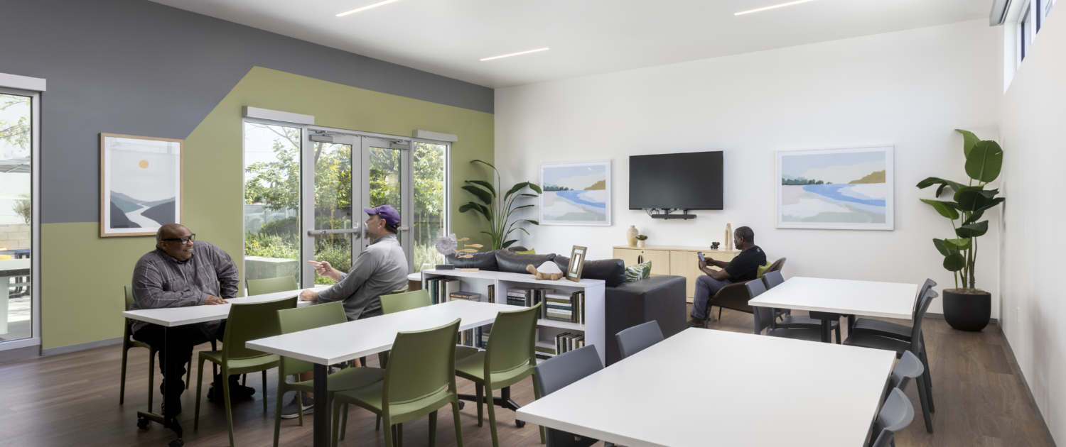 Community gathering space with relaxing abstract art. Looking into space with several modular white tables, green tables, TV in background, and residents chatting.