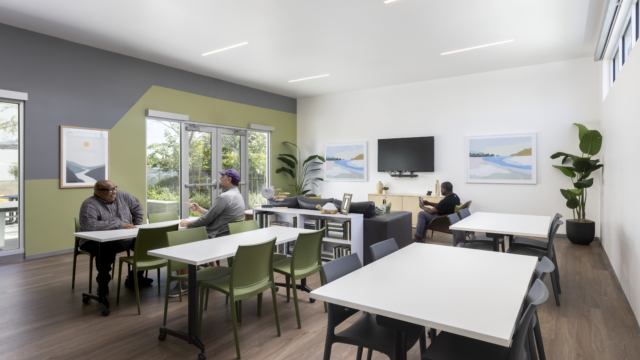 Community gathering space with relaxing abstract art. Looking into space with several modular white tables, green tables, TV in background, and residents chatting.