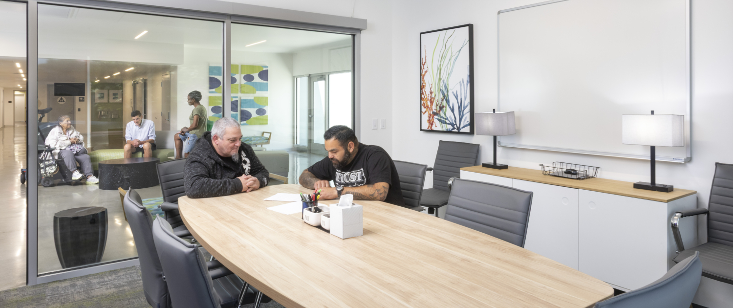 Flex-use small conference room with two residents sitting at wooden table with grey chairs. Behind them is the office glass wall with a view on what's happening in the lobby. There, residents are connecting.