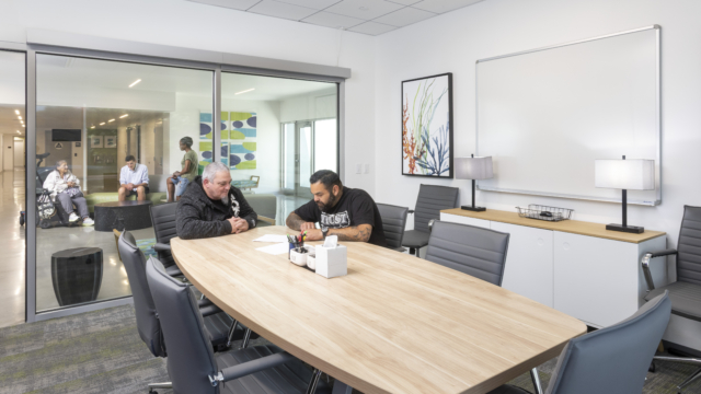 Flex-use small conference room with two residents sitting at wooden table with grey chairs. Behind them is the office glass wall with a view on what's happening in the lobby. There, residents are connecting.