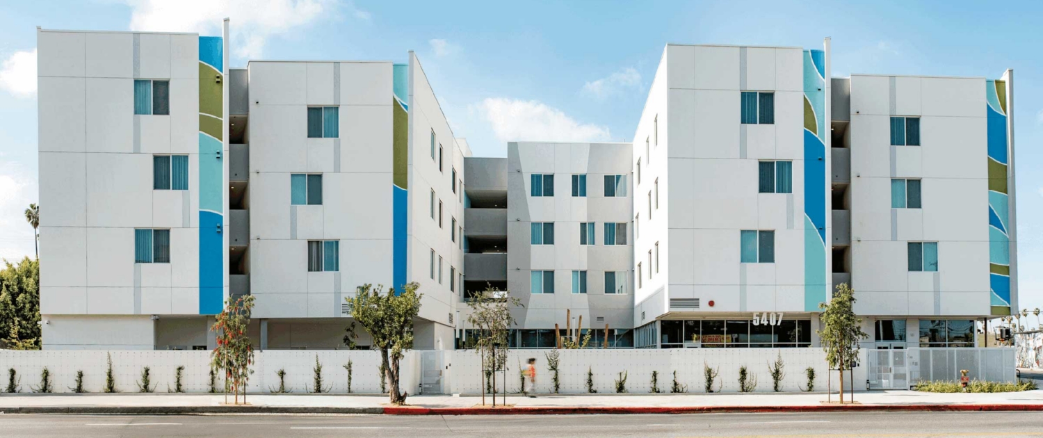 A modern, four-story apartment building with a white facade and blue and green accent panels, featuring balconies, large windows, and a landscaped area in front along a wide street.