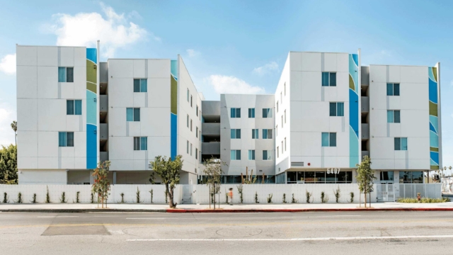 A modern, four-story apartment building with a white facade and blue and green accent panels, featuring balconies, large windows, and a landscaped area in front along a wide street.