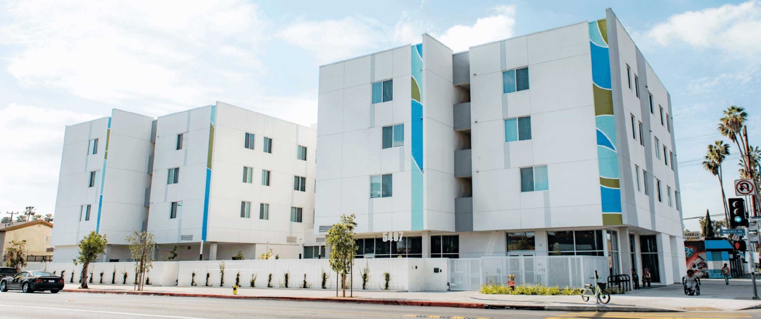 A modern, white apartment building with colorful vertical accents stands along a street with a yellow-striped crosswalk, palm trees, and clear blue sky in the background.