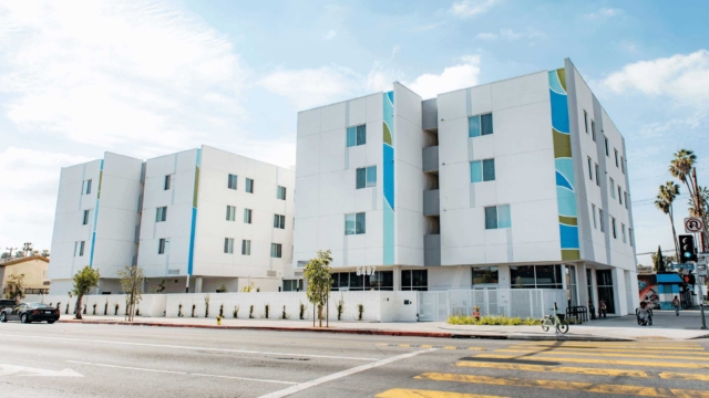 A modern, white apartment building with colorful vertical accents stands along a street with a yellow-striped crosswalk, palm trees, and clear blue sky in the background.