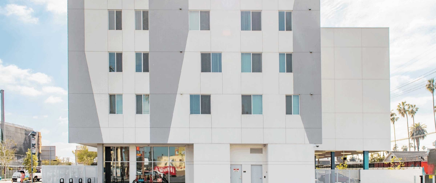 A modern, four-story white building with geometric gray accents on the facade, large windows on the ground floor, and smaller windows above, set against a sunny sky with palm trees visible in the background.