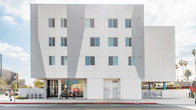 A modern, four-story white building with geometric gray accents on the facade, large windows on the ground floor, and smaller windows above, set against a sunny sky with palm trees visible in the background.