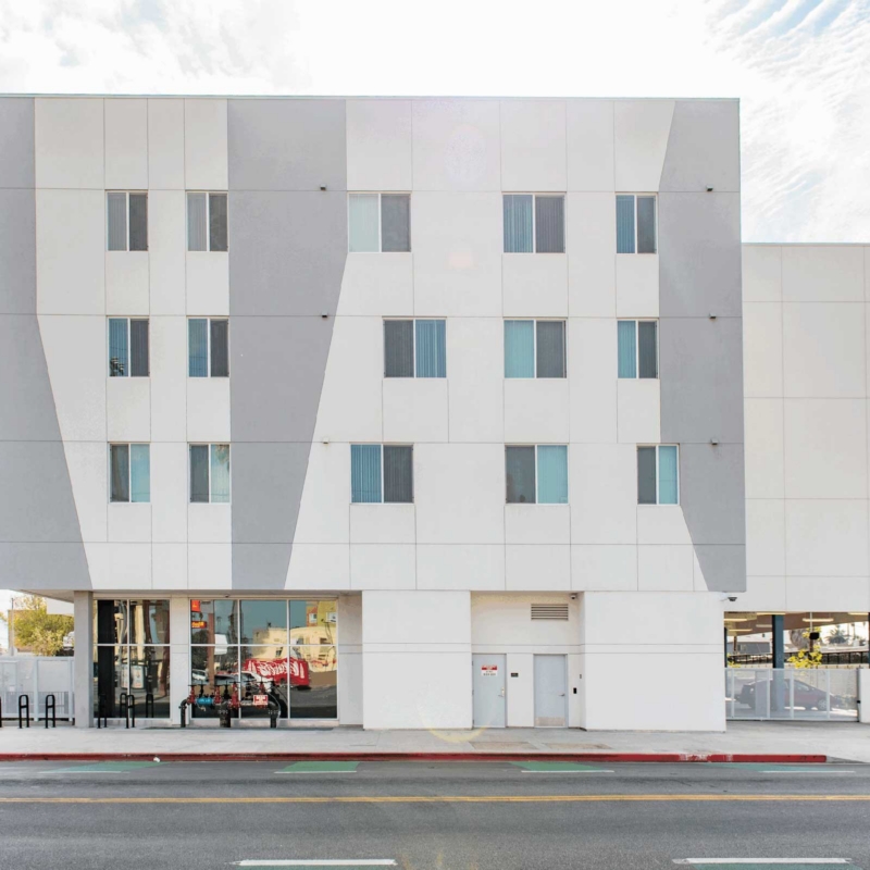A modern, four-story white building with geometric gray accents on the facade, large windows on the ground floor, and smaller windows above, set against a sunny sky with palm trees visible in the background.