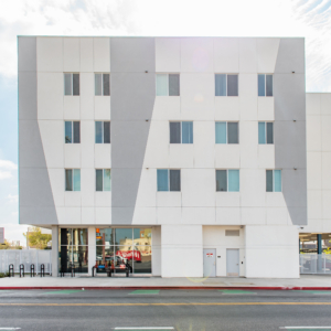 A modern white housing building with geometric gray accents, large windows on the ground floor, smaller windows above, and bicycle racks near the entrance—an inviting spot to find housing, viewed from across the street.