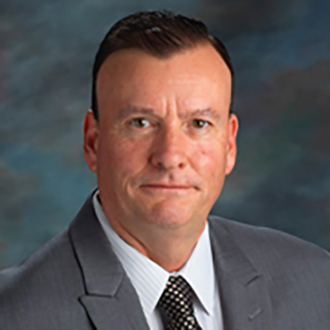 A man in a gray suit, white shirt, and patterned tie poses for a formal portrait against a blurred dark background. He has short dark hair and a serious expression.