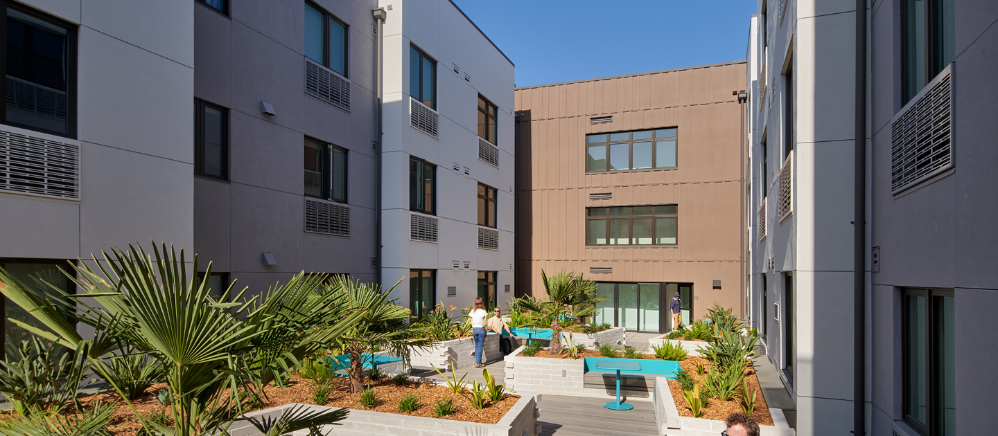 A landscaped courtyard between modern apartment buildings with people sitting and talking at blue tables amid green plants under a clear sky.