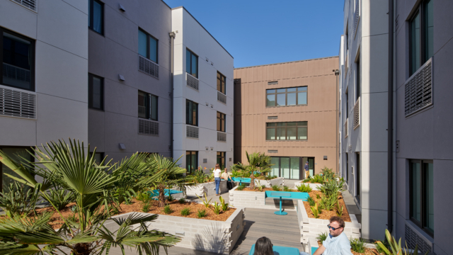 A landscaped courtyard between modern apartment buildings with people sitting and talking at blue tables amid green plants under a clear sky.