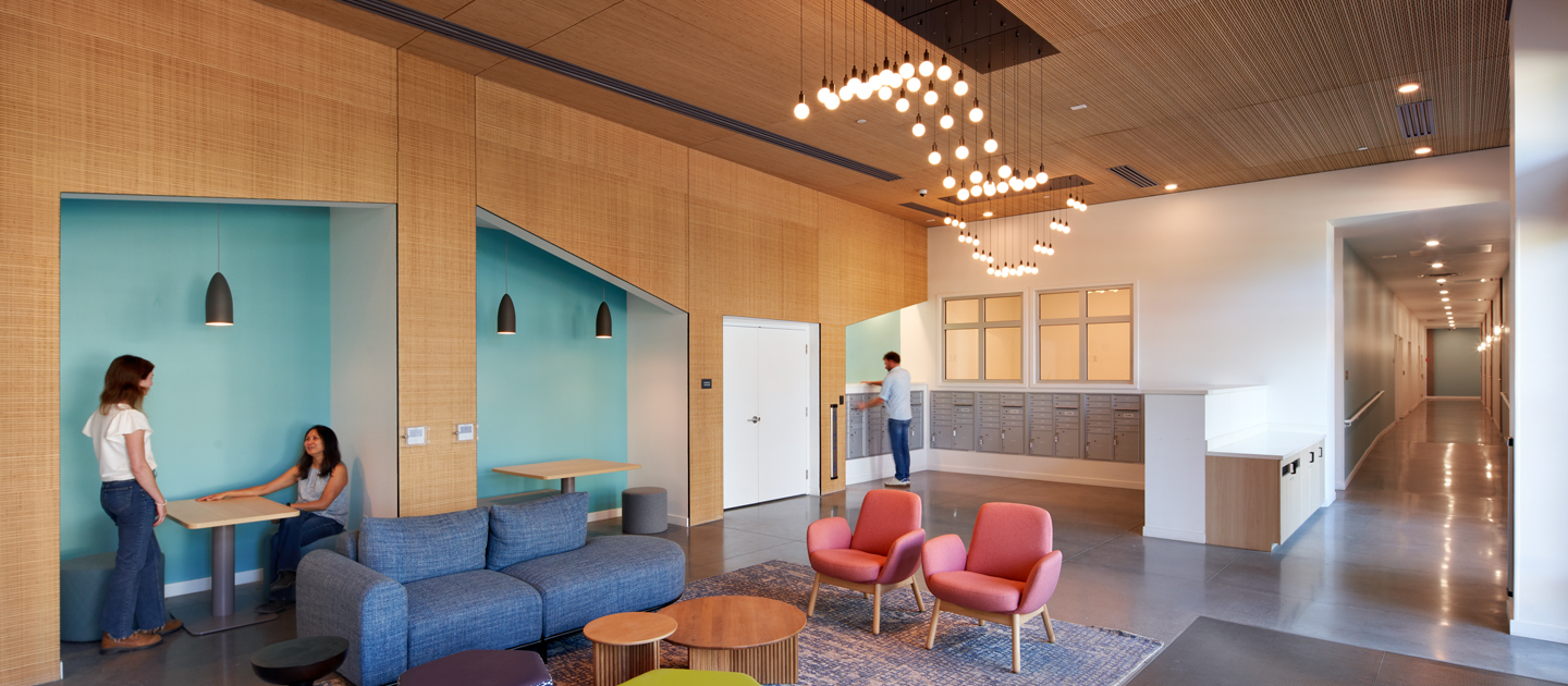 A modern office lounge with colorful seating, a blue rug, hanging lights, and wood accents. Two people talk at a booth, and another person stands near a kitchenette by a hallway.