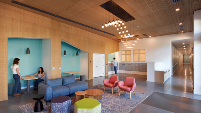 A modern office lounge with colorful seating, a blue rug, hanging lights, and wood accents. Two people talk at a booth, and another person stands near a kitchenette by a hallway.