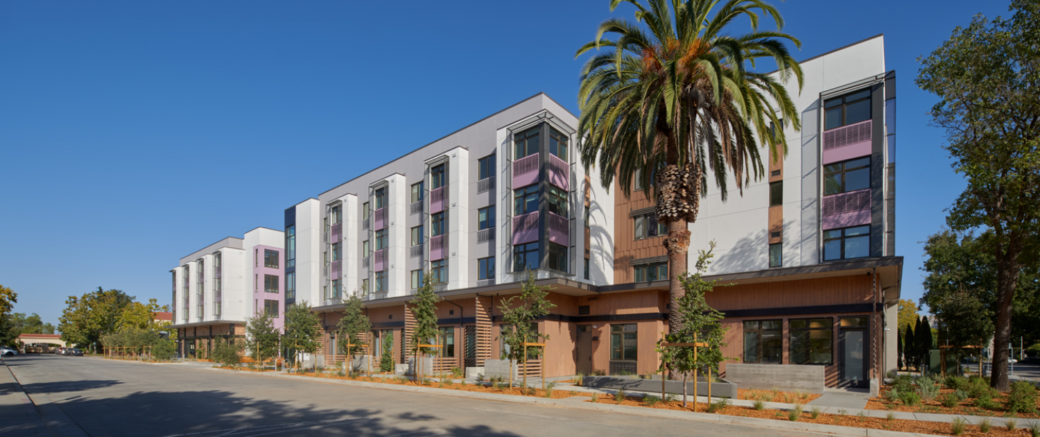 A modern three-story apartment building with large windows, balconies, and pink accents stands along a quiet street lined with small trees and a tall palm tree under a clear blue sky.