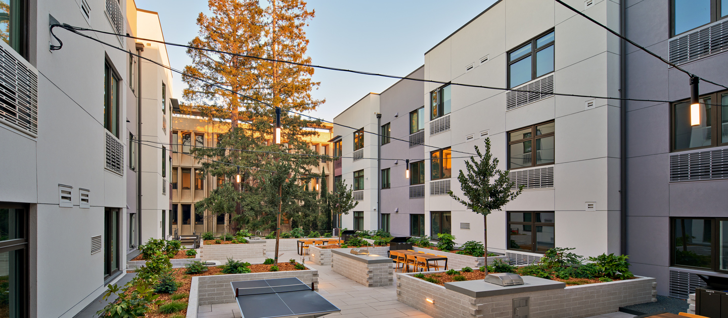 Modern apartment courtyard with table tennis, outdoor seating, string lights, and planters, surrounded by white buildings and tall trees in the background under a clear sky.