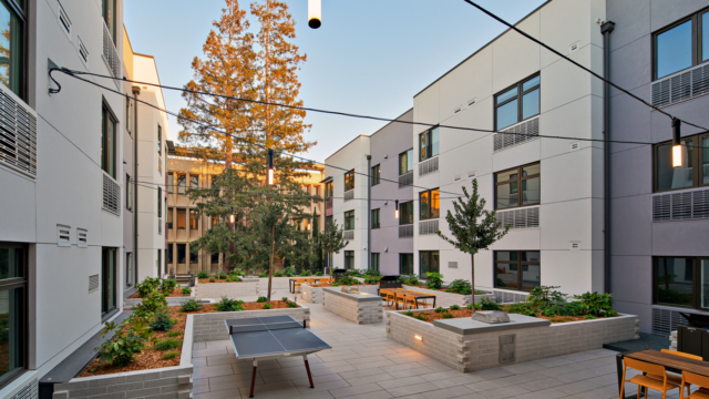 Modern apartment courtyard with table tennis, outdoor seating, string lights, and planters, surrounded by white buildings and tall trees in the background under a clear sky.