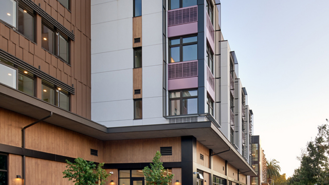 Modern multi-story apartment building with large windows and balconies, people sitting at outdoor tables, and others walking along a wide sidewalk lined with small trees and landscaping.