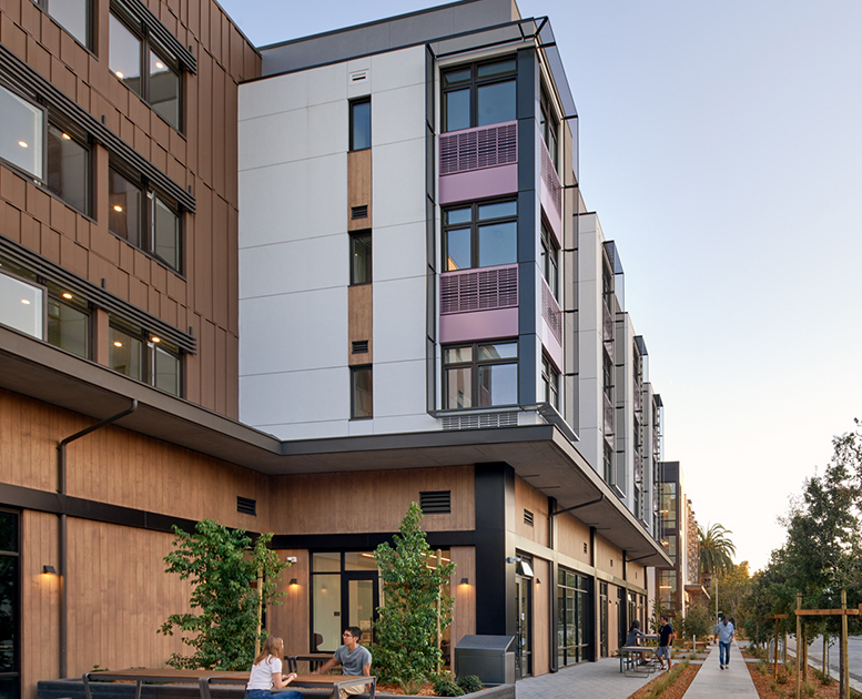Modern multi-story apartment building with large windows and balconies, people sitting at outdoor tables, and others walking along a wide sidewalk lined with small trees and landscaping.