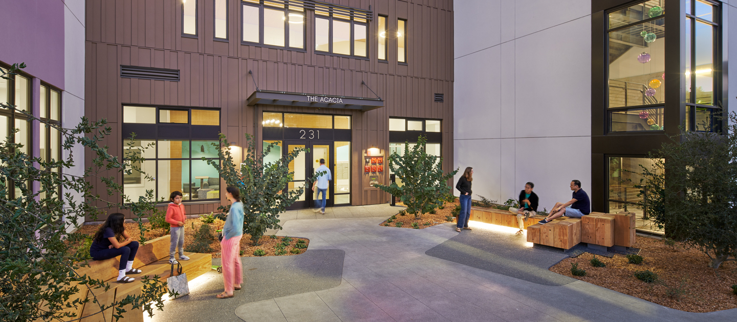 People sit and stand on benches in a well-lit modern courtyard outside a building entrance with large windows and plants; some are talking or using their phones, and indoor lights reveal colorful decorations.