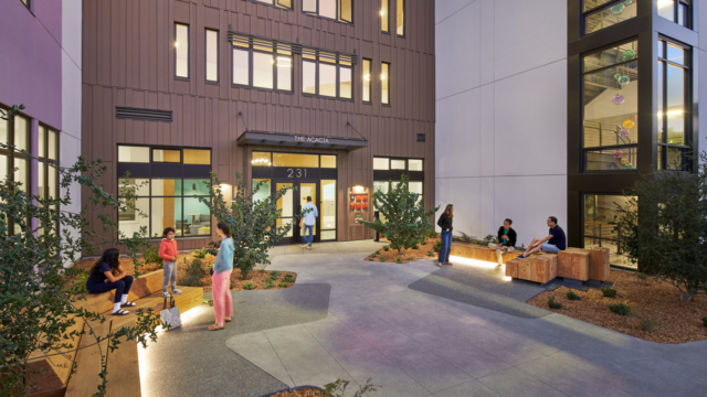People sit and stand on benches in a well-lit modern courtyard outside a building entrance with large windows and plants; some are talking or using their phones, and indoor lights reveal colorful decorations.