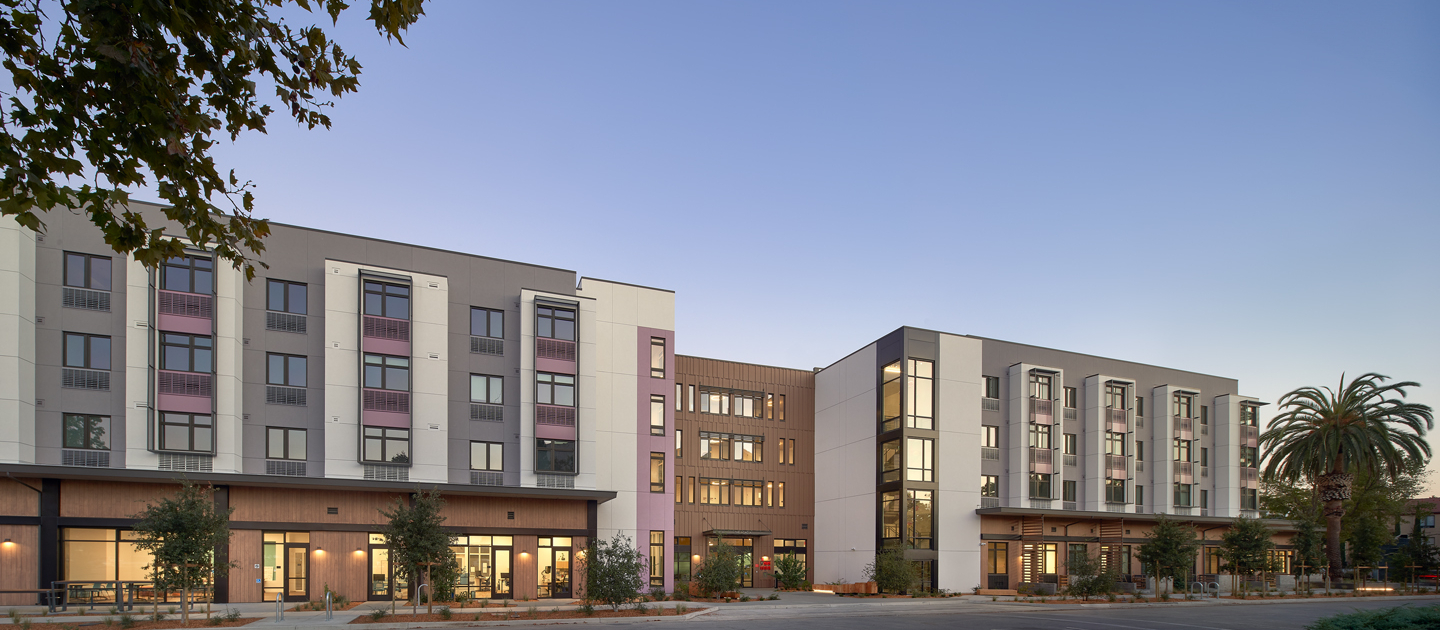 A modern multi-story apartment building with large windows, balconies, and ground-floor entrances, surrounded by young trees and a nearly empty parking area at sunset.