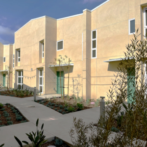 A modern housing apartment building with beige walls, green doors, and tall windows, surrounded by landscaped walkways and desert plants under a partly cloudy sky.