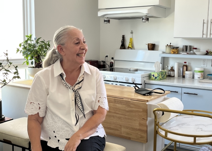 Photo Aug 26 2025, 11 51 16 AM An older woman with gray hair, wearing a white blouse and tie, sits smiling on a chair in a bright Harrower Village kitchen with white cabinets, plants, and various kitchen items on the counters.