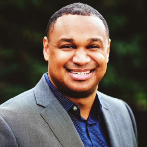 A man in a gray suit and navy shirt smiles at the camera, standing outdoors with a blurred green background.