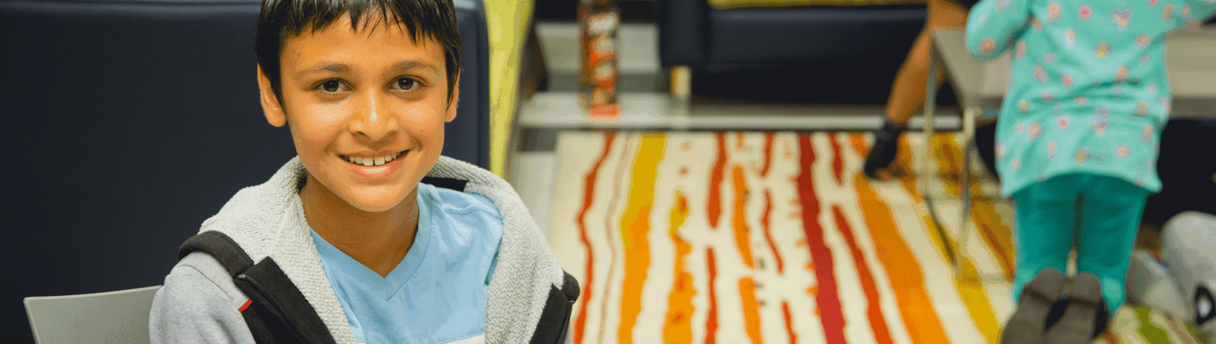 A young boy with short dark hair sits and smiles at the camera in a colorful room with striped carpet. In the background, two children play on the floor near a table and couch.
