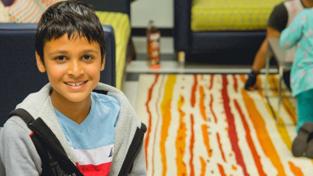 A smiling boy with short dark hair sits indoors on a chair, wearing a light blue and white shirt with a gray jacket. Behind him, children play on a rug with yellow and red stripes in a cozy room.
