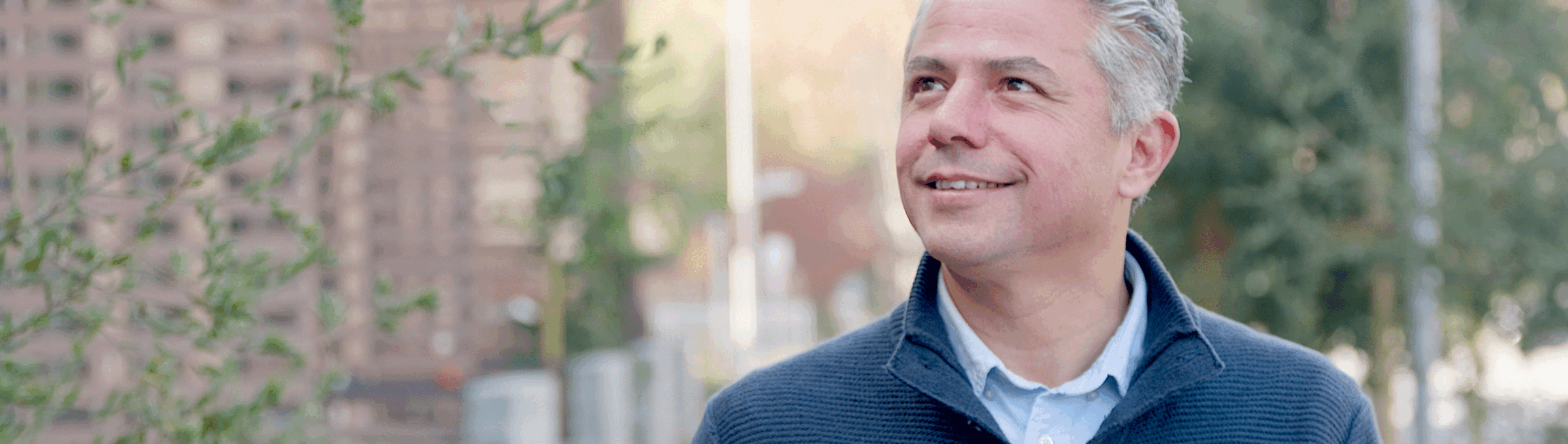 A middle-aged man with gray hair, wearing a blue sweater over a collared shirt, stands outdoors, smiling and looking up, with trees and wooden structures in the blurred background.