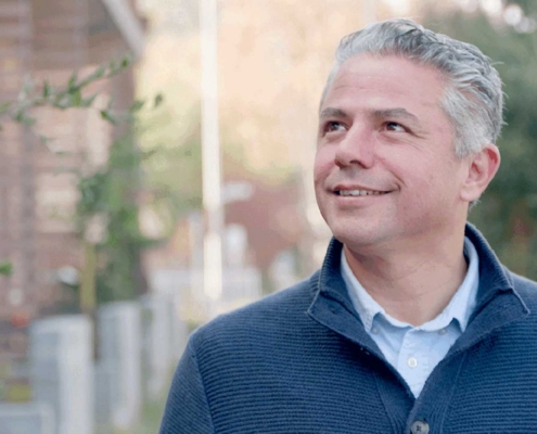 A man with gray hair wearing a blue sweater and light blue shirt stands outdoors, smiling and looking up. Trees and a blurred building are in the background on a sunny day.