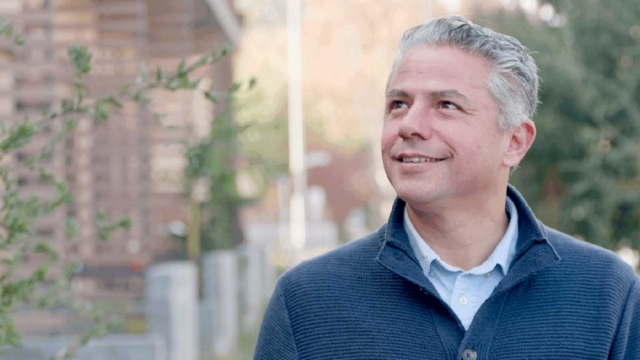 A man with gray hair wearing a blue sweater and light blue shirt stands outdoors, smiling and looking up. Trees and a blurred building are in the background on a sunny day.