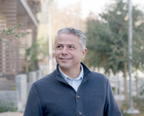 A middle-aged man with gray hair smiles while standing outdoors. He is wearing a textured gray sweater over a collared shirt, with trees and a modern building in the blurred background.