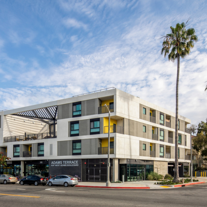 Four-story building with open air courtyard on upper floor. Building is white with blue window reflections and accents of yellow. Ground floor has floor-to-ceiling glass on corner, lush landscaping, and the facade is board-form concrete spanning the length of the building to the right of the storefront glass.